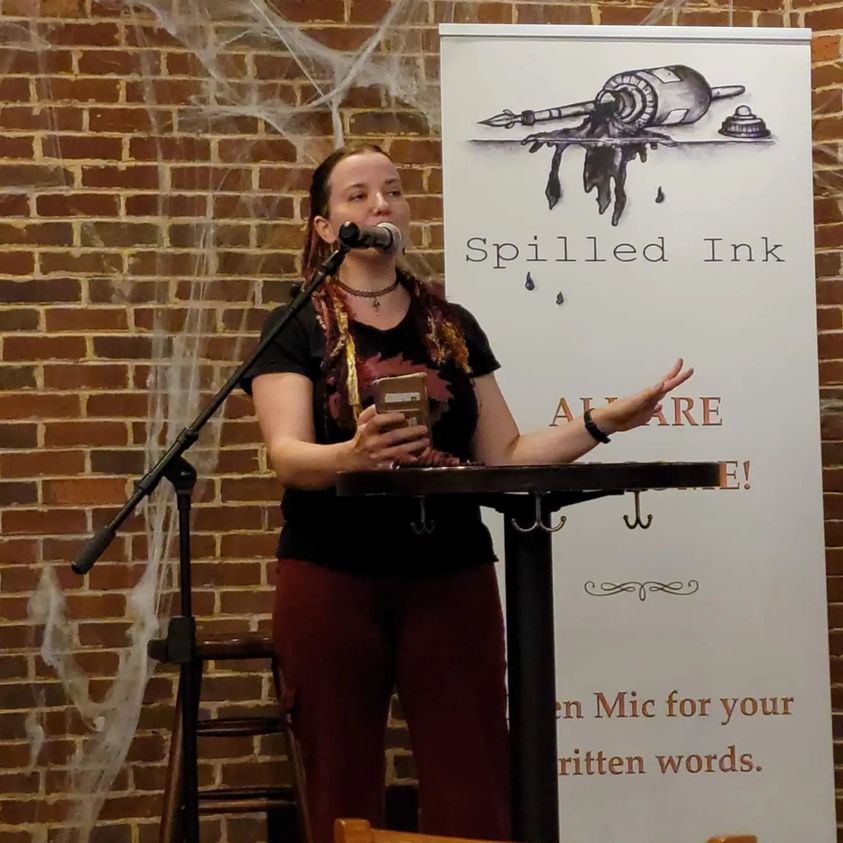 Morgan at the mic, rocking some red&gold yarn falls, with the Spilled Ink banner behind her. "Open mic for your written words."